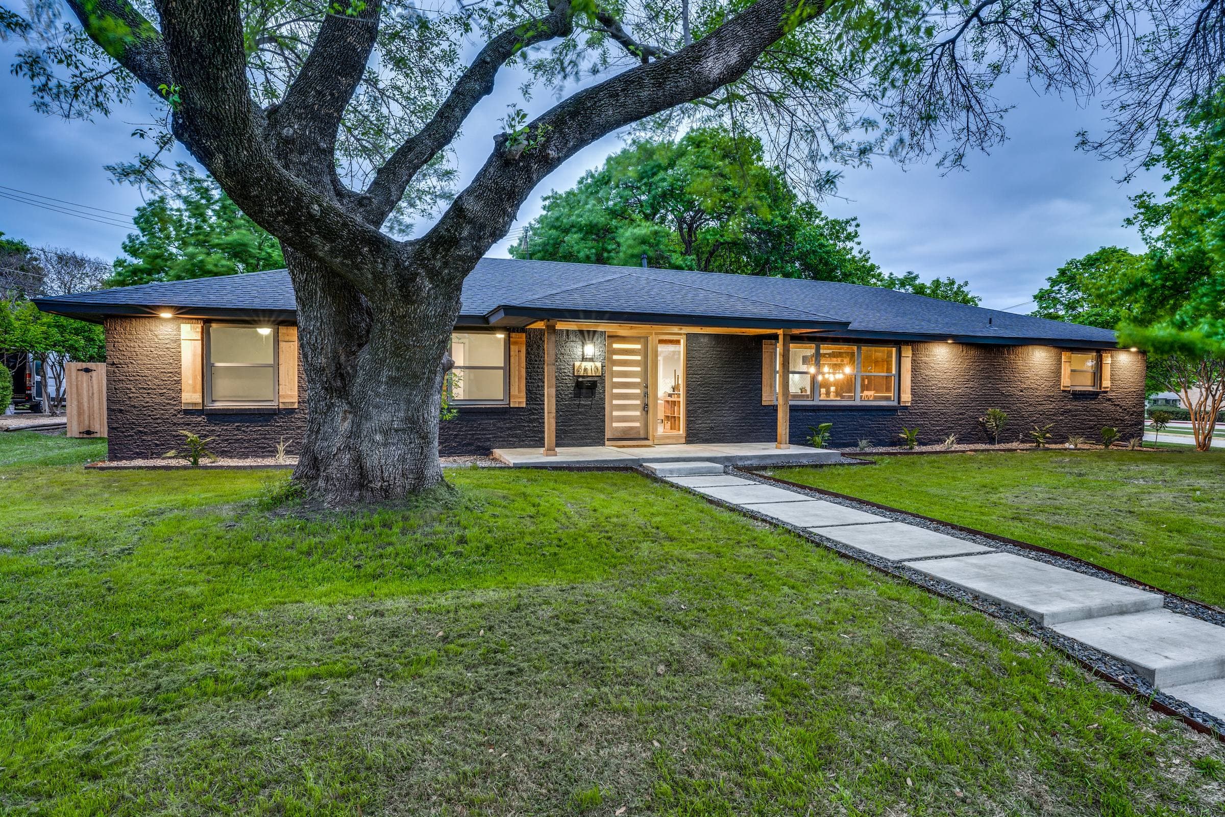 Single-story dark brick home with a covered entry and warm exterior lighting at dusk